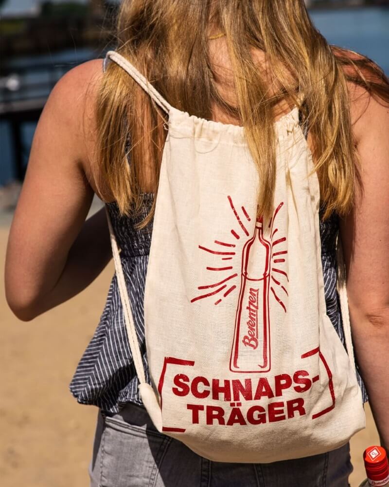 Berentzen Schnapstraeger Frau trägt Creme farbigen Jute Beutel mitrotem Berentzen Flasche Logo und Schriftzug: Schnapsträger